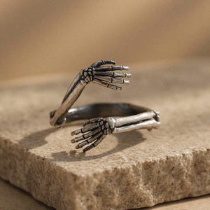 Puede incluir: Un anillo plateado con forma de dos manos de esqueleto extendidas. El anillo está sobre una superficie texturizada de color marrón claro. El diseño del anillo presenta dedos y brazos esqueléticos detallados.