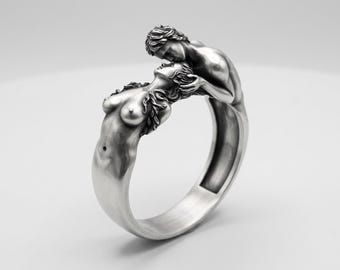 Könnte beinhalten: Ein silberfarbener Ring mit einem skulpturalen Design aus zwei sich umarmenden Figuren. Die Ringschiene wird durch die Körper der Figuren gebildet, wobei sich eine Figur zurücklehnt, während die andere sich darüber beugt.