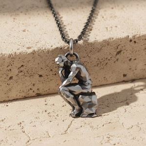 Puede incluir: Collar con colgante plateado que presenta una escultura geométrica de una persona sentada en una pose contemplativa. El colgante cuelga de una cadena oscura. La escultura está detallada con facetas angulares.