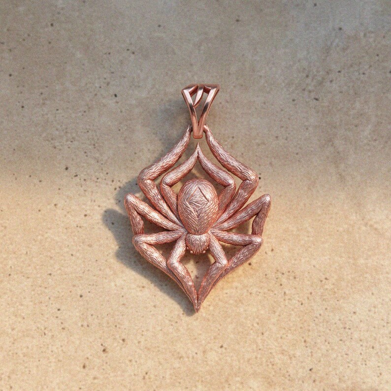 May include: A rose gold spider pendant with intricate detailing. The pendant features a stylized spider design with eight legs and a textured body. The bail is a simple loop for a chain.
