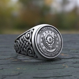Op de afbeelding: Zilverkleurige ring met het embleem van het Criminal Investigation Command van het Amerikaanse leger. De ring heeft de woorden "United States Army" en "Criminal Investigation Command" rond een centraal embleem. De zijkanten van de ring zijn versierd met gedetailleerde patronen.
