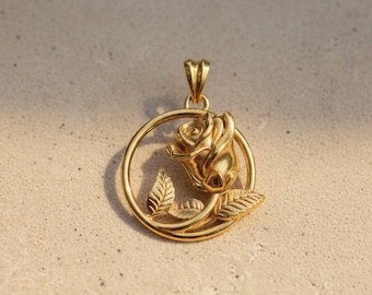 May include: A gold-toned metal pendant featuring a circular frame enclosing a three-dimensional rose flower with two detailed leaves. The pendant includes a small triangular bail at the top for attachment to a chain, showcasing a polished finish.