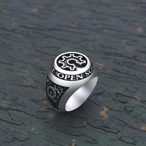 May include: A silver-toned ring featuring a circular top with a gear icon and the text "OPEN SOURCE" engraved along the border. The sides of the ring display an intricate, textured pattern with additional small gear motifs integrated into the design.