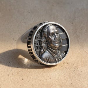 Könnte beinhalten: Ein silberner Ring mit einem detaillierten Relief von Benjamin Franklin, ähnlich einer 100-Dollar-Note. Der Ring hat ein breites Band mit der Gravur "THE UNITED" um die Lünette und ist vor einem neutralen Hintergrund platziert.