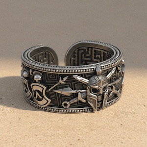 May include: Silver-toned adjustable ring featuring a detailed warrior design. The ring showcases a helmet, spears, and shield motifs, with a textured band and intricate patterns. A statement piece for those interested in historical or fantasy themes.