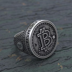 Könnte beinhalten: Ein silberner Ring mit einem Bitcoin-Symbol auf der Vorderseite. Der Ring ist mit den Worten "Monetary Metal" und einer Reihe von Binärcode graviert.