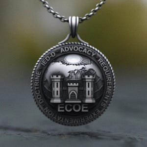 May include: Silver pendant necklace with a circular medallion. The medallion features a castle design at the centre, with text around the edge including "IED-D ADVOCACY REQUIREMENTS" and "ECOE". The necklace is suspended from a silver chain.