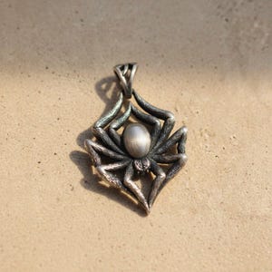 May include: A silver-colored spider pendant with intricate detailing. The pendant features a central oval body and eight legs, suspended from a loop at the top. The spider is set against a neutral background.