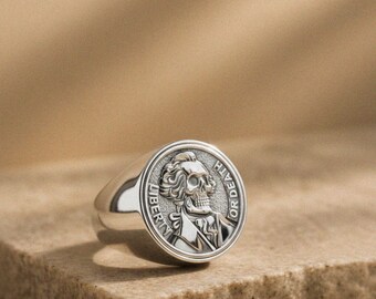 May include: A silver signet ring with a detailed skull design. The ring features the words "LIBERTY" and "OR DEATH" around the skull. The ring is on a stone surface.