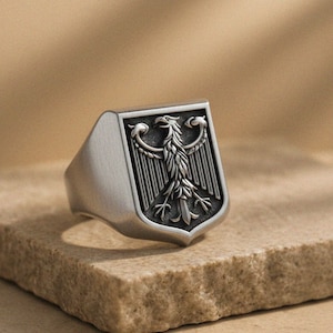 Puede incluir: Un anillo plateado con una cara en forma de escudo. La cara presenta un relieve detallado de un águila con las alas extendidas. El anillo está sobre una superficie texturizada de color marrón claro.