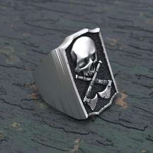 May include: A silver ring featuring a skull and crossed axes design. The ring is displayed on a dark green wooden surface.