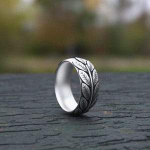 Può includere: Un anello in metallo color argento caratterizzato da un motivo in rilievo continuo di foglie dettagliate che avvolgono la fascia esterna. L'interno dell'anello è liscio e lucido, mentre la superficie esterna presenta una finitura scura ossidata.