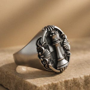 May include: A silver-toned ring featuring a detailed chess queen design. The ring has an oval face with a checkered background and ornate floral-like embellishments. The queen piece is prominently displayed in the center.