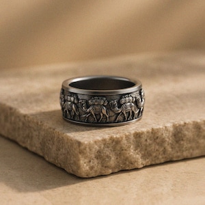 May include: A silver-toned ring with a detailed relief of camels. The ring has a wide band with the camel design wrapping around the circumference. The ring is displayed on a textured, light brown surface.