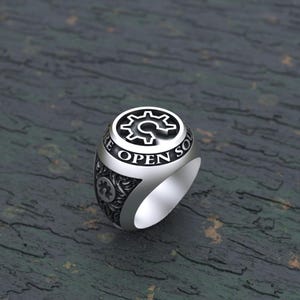 以下が含まれることがあります： 歯車模様の黒いデザインと、歯車の周りに「We Are Open Source」という文字が刻まれた銀の指輪。指輪の側面には、細かなデザインが施されています。