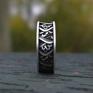May include: A silver ring with a detailed wave pattern. The ring has a dark, textured background with a repeating design of stylized waves and dots. The edges of the ring are polished silver.