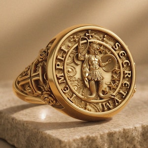 Könnte beinhalten: Ein goldener Siegelring mit einem zentralen Relief einer Figur mit Schlangenschwanz, die eine Peitsche und einen Schild hält. Die kreisförmige Fläche trägt die Inschrift TEMPLI SECRETVM und ist von Sternen umgeben. Der Ring zeigt florale Schnitzereien.