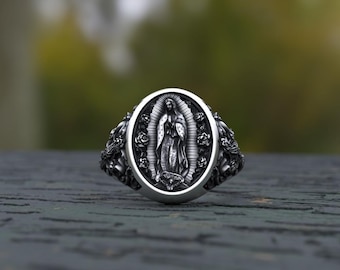 May include: A silver-toned metal ring featuring an oval centerpiece with a relief depiction of the Virgin of Guadalupe surrounded by roses. The band is detailed with ornate floral carvings on both sides, showcasing a dark, oxidized finish.