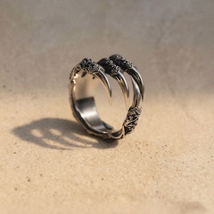 Könnte beinhalten: Ein silberner Ring mit einem Krallendesign, das drei scharfe, gebogene Krallen aufweist. Der Ring hat aufwendige Details am Band, und die Krallen sind deutlich sichtbar. Der Ring ist vor einem neutralen Hintergrund platziert.