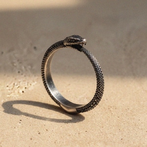 May include: A silver-toned ring shaped like a snake, with the snake's head at the top. The snake's body forms the ring's band. The ring is on a light brown surface.