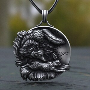 May include: A circular silver-toned pendant featuring a detailed relief carving of a wolf leaning over a rabbit. The pendant has a dark, oxidized finish to highlight the texture of the fur and the woodland scene, suspended from a silver chain.