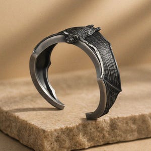 Puede incluir: Un brazalete de metal en tono plateado oscuro con un diseño de murciélago esculpido. El murciélago está posicionado en la parte superior central con sus alas extendiéndose a lo largo de los lados de la banda, mostrando texturas detalladas.