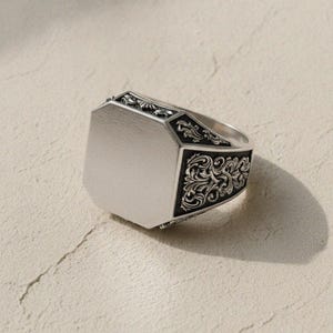May include: A silver signet ring featuring a smooth, flat, octagonal top surface. The sides of the ring are decorated with intricate, dark, recessed scrollwork patterns that contrast with the polished, bright silver finish of the band.