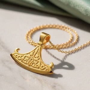 May include: A gold-colored pendant necklace featuring a stylized axe design. The pendant has an ornate, engraved pattern and is suspended from a delicate chain. The necklace is displayed on a marble surface with a soft green fabric in the background.