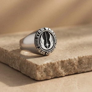 May include: A silver ring with a circular emblem featuring the words "STAR WARS JEDI ORDER" around a central design. The ring is set on a textured, light brown surface.
