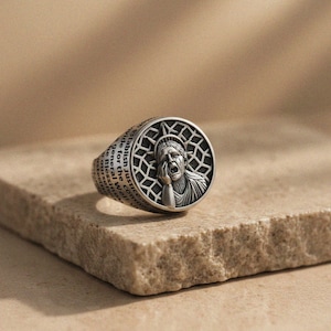 Könnte beinhalten: Ein silberner Ring mit einem Design der leidenden Freiheitsstatue. Die Ringoberfläche hat einen kreisförmigen Rahmen mit einem geometrischen Muster. Das Ringband ist mit Text graviert. Der Ring wird auf einer strukturierten, hellbraunen Oberfläche präsentiert.