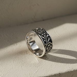 Può includere: Un anello d'argento con un motivo floreale. L'anello presenta un design dettagliato di fiori e foglie attorno alla fascia. L'anello è esposto su una superficie neutra, con ombre che aggiungono profondità.