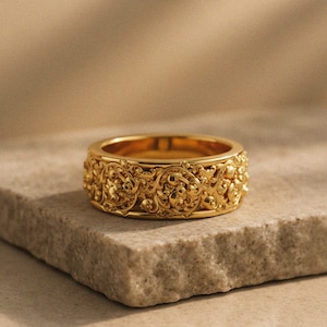 Puede incluir: Un anillo dorado con un diseño floral ornamentado y detallado. El anillo está colocado sobre una superficie de piedra, mostrando sus intrincados grabados y su estética general. El diseño del anillo presenta un patrón repetido de flores y hojas.