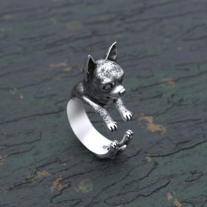 Könnte beinhalten: Ein silberner Ring mit einer detaillierten Chihuahua-Skulptur. Der Chihuahua ist in einer verspielten Pose dargestellt, mit erhobenen Pfoten und seitlich geneigtem Kopf. Der Ring ist aus hochwertigem Metall gefertigt und ein einzigartiges und stilvolles Accessoire.