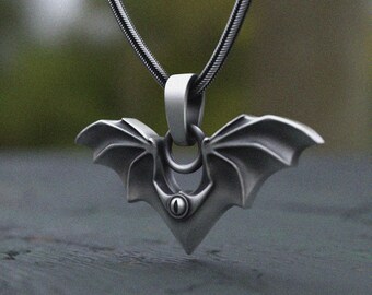 May include: A dark gray metal pendant shaped like a bat with outstretched wings, featuring a central eye detail. The pendant hangs from a matching dark gray snake chain necklace, showcasing a smooth, matte finish and intricate wing structure.