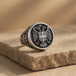 May include: Silver ring with a detailed double-headed eagle emblem and shield. The ring has an engraved band design and sits on a textured, light brown surface. A piece of jewellery with a historical theme.