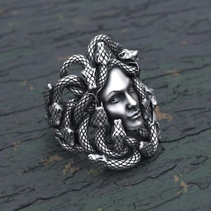 Può includere: Un anello d'argento con una scultura dettagliata di Medusa, la figura mitologica greca con i capelli di serpenti. L'anello è un pezzo di grande impatto con dettagli intricati e un design accattivante.