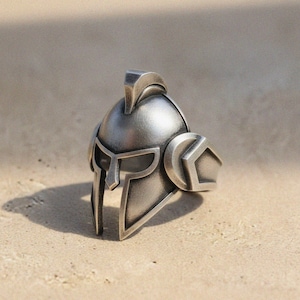 May include: A silver-toned ring in the shape of a Spartan helmet. The ring showcases intricate details, such as the helmet's crest, face guard, and side shields. The ring is crafted from a metallic material and is presented on a light-coloured surface.