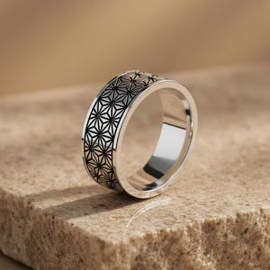 May include: A silver ring with a geometric black pattern. The ring is set on a textured, light brown surface. The ring's design features repeating star-like shapes around the band.