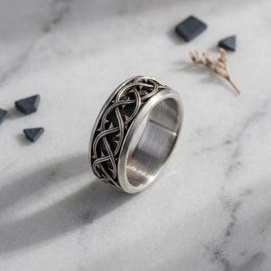 May include: A silver-toned metal ring featuring an intricate, raised interwoven knotwork pattern around the exterior band. The band has a smooth, polished interior surface and a dark, recessed background within the detailed outer design.