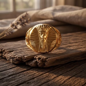 Può includere: Un anello color oro caratterizzato da un rilievo dettagliato e rialzato del volto di un faraone egizio centrato su una piastra circolare. Il design include intricate linee radianti che circondano la testa, con motivi decorativi sulla fascia.