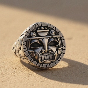 Puede incluir: Anillo plateado con un diseño detallado de máscara tiki. El anillo tiene una banda texturizada y una cara con una expresión de mueca. El anillo está sobre un fondo arenoso.