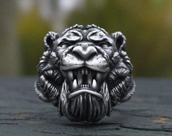 May include: A silver-toned ring featuring a detailed, three-dimensional tiger head design. The tiger is depicted with an open mouth, bared teeth, and textured fur detailing across the face and mane, finished with dark oxidised accents.