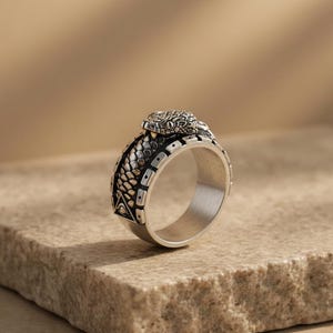 Op de afbeelding: Een zilveren ring met een gedetailleerd slangenontwerp. De ring heeft een slangenkop aan de bovenkant en ingewikkelde schubpatronen langs de band. De ring wordt gepresenteerd op een getextureerd, lichtbruin oppervlak.