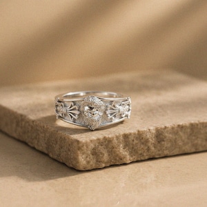 Könnte beinhalten: Ein silberner Ring mit einem zentralen Relief eines bärtigen Gesichts mit detailliertem Haar und Gesichtszügen, flankiert von symmetrischen, ornamentalen, blumenartigen Motiven auf beiden Seiten des Rings, mit polierter Oberfläche.