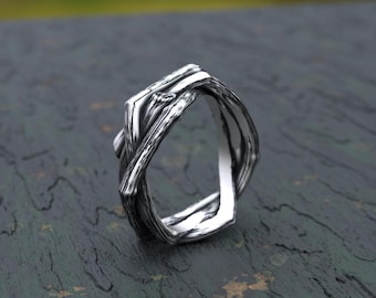 Könnte beinhalten: Ein silberner Ring mit einer texturierten, organischen Oberfläche, die wie ineinander verschlungene Zweige oder Äste aussieht. Das Metall hat ein poliertes Finish mit dunklen, vertieften Bereichen, die die unregelmäßigen Linien betonen.