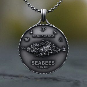 May include: Silver pendant necklace with a detailed design. The pendant features the text "WE BUILD, WE FIGHT" and "SEABEES 'CAN DO!'" with a bee-like emblem. The necklace has a chain with a lobster clasp.