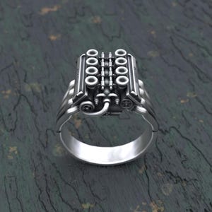 Puede incluir: Un anillo de plata con un diseño de motor detallado. El anillo presenta un bloque de motor estilizado con múltiples cilindros y un colector de escape detallado.