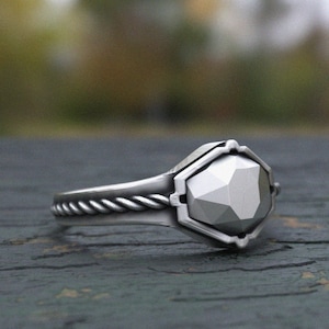 Könnte beinhalten: Ein silberner Ring mit einem geometrischen, facettierten grauen Stein. Der Ring hat ein gedrehtes Seil-Design am Band und eine sechseckige Fassung um den Stein. Der Ring ist vor einem unscharfen Hintergrund.
