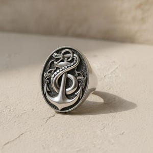 May include: A silver-toned ring featuring an oval face with a raised relief design of a nautical anchor entwined with a serpent. The background of the oval is textured and dark, providing contrast to the polished, raised metal elements of the ring.