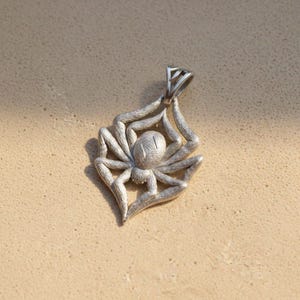 May include: A silver spider pendant with intricate detailing. The pendant features a detailed spider design with eight legs and a textured body. The bail is a triangular shape. The pendant is likely made of metal.